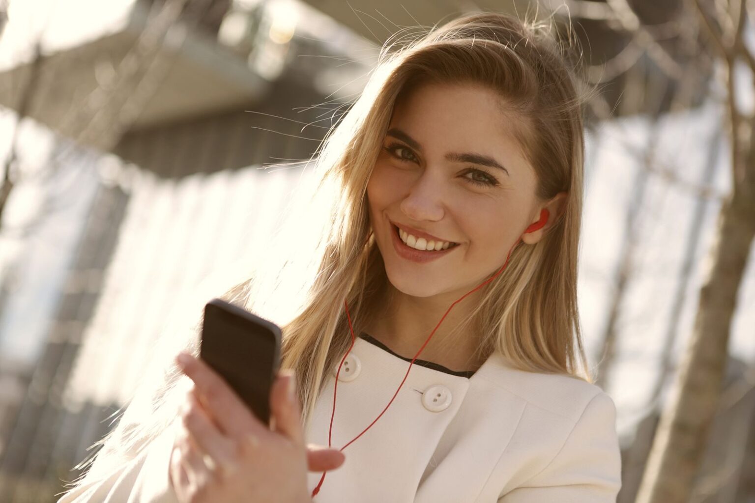 A smiling woman with headphones and smartphone enjoying music outdoors on a sunny day.