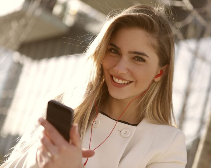 A smiling woman with headphones and smartphone enjoying music outdoors on a sunny day.