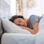 A serene woman sleeps comfortably in bed, surrounded by soft white linens in a sunlit room.
