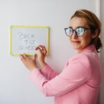 A cheerful female teacher in a pink blazer writes 'Back to School' on a whiteboard.