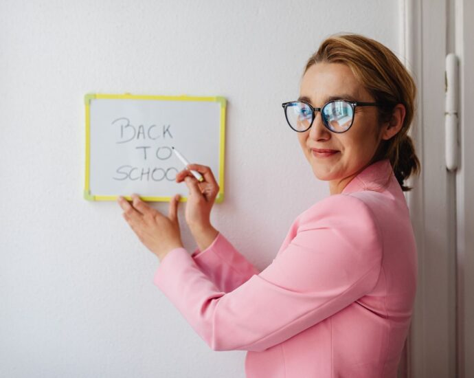 A cheerful female teacher in a pink blazer writes 'Back to School' on a whiteboard.