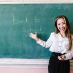 Smiling teacher in a classroom pointing at back to school message on a chalkboard.