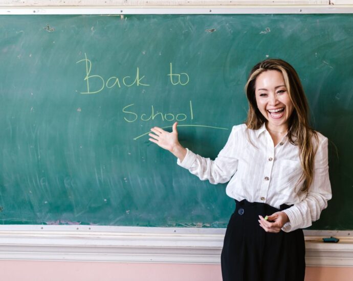 Smiling teacher in a classroom pointing at back to school message on a chalkboard.