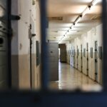 A long prison corridor with rows of metal doors viewed through dark bars