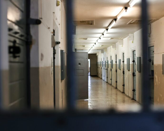 A long prison corridor with rows of metal doors viewed through dark bars