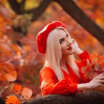 Blonde woman in red beret surrounded by vibrant autumn leaves in a forest setting.