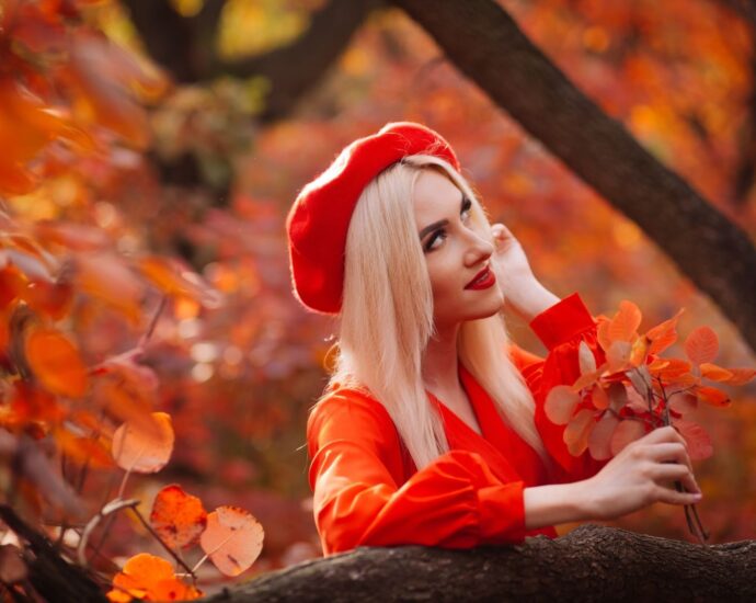 Blonde woman in red beret surrounded by vibrant autumn leaves in a forest setting.